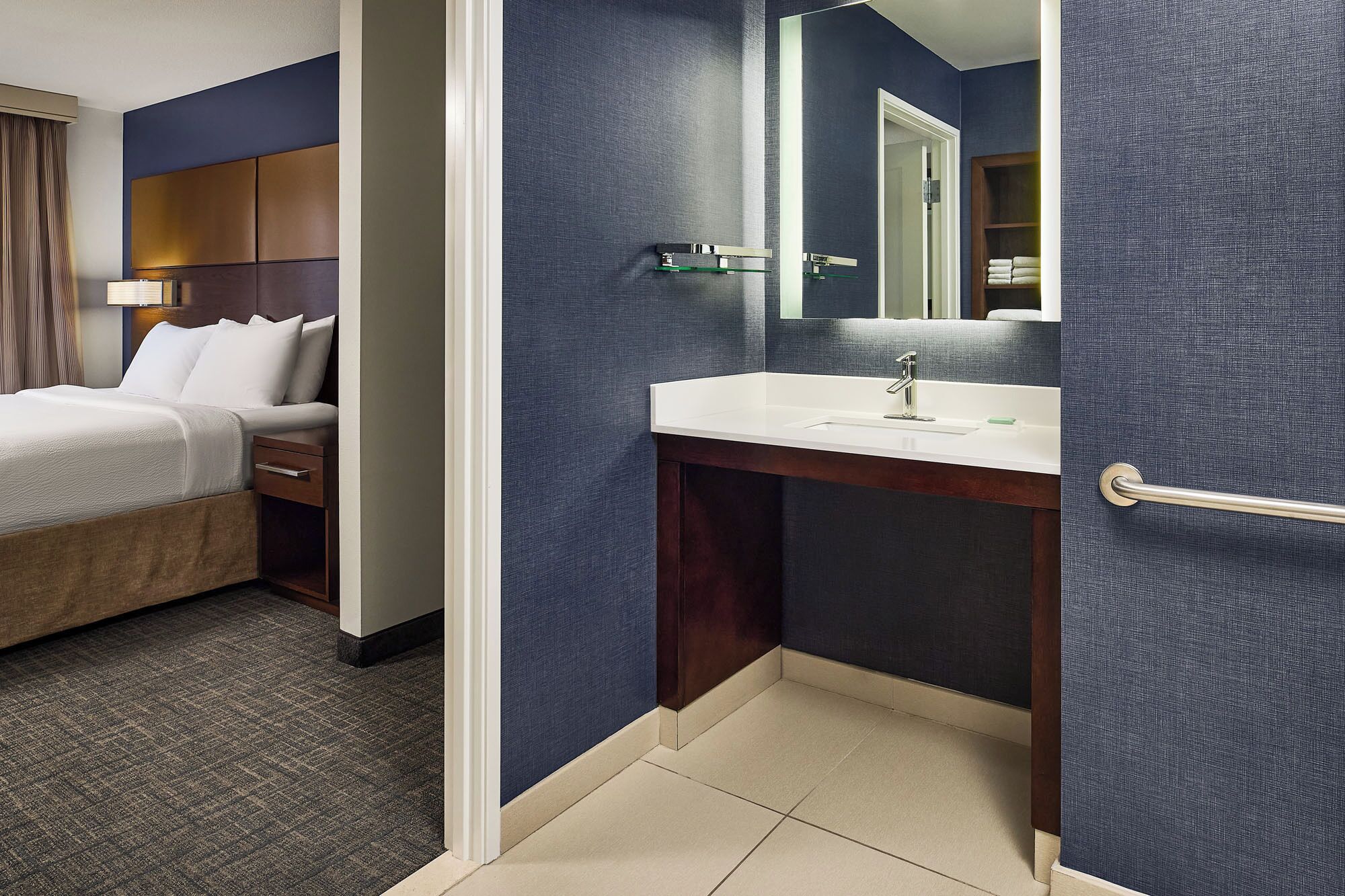 Una habitación de hotel con cama, baño con lavamanos y espejo, y una puerta que da a otra habitación.