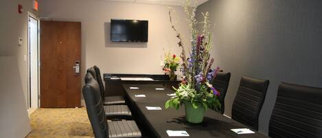 Uma sala de conferências com uma mesa comprida, cadeiras, uma televisão na parede e um arranjo floral.