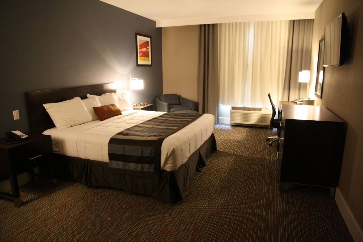 A hotel room with a bed, bedside table, chair, desk, and a large window with curtains.