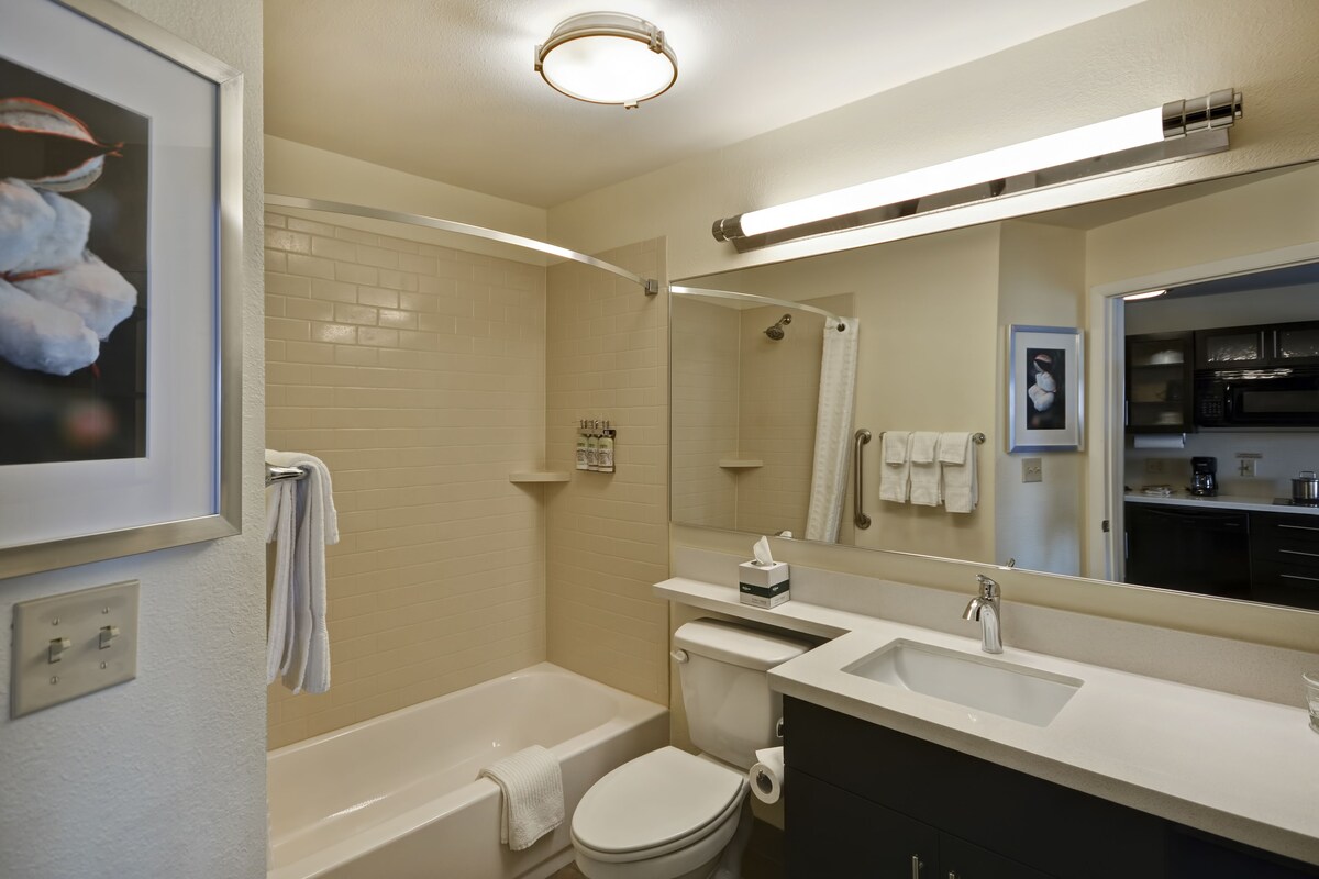 Un baño moderno con una bañera blanca, inodoro y lavamanos, un espejo grande y un cuadro en la pared.