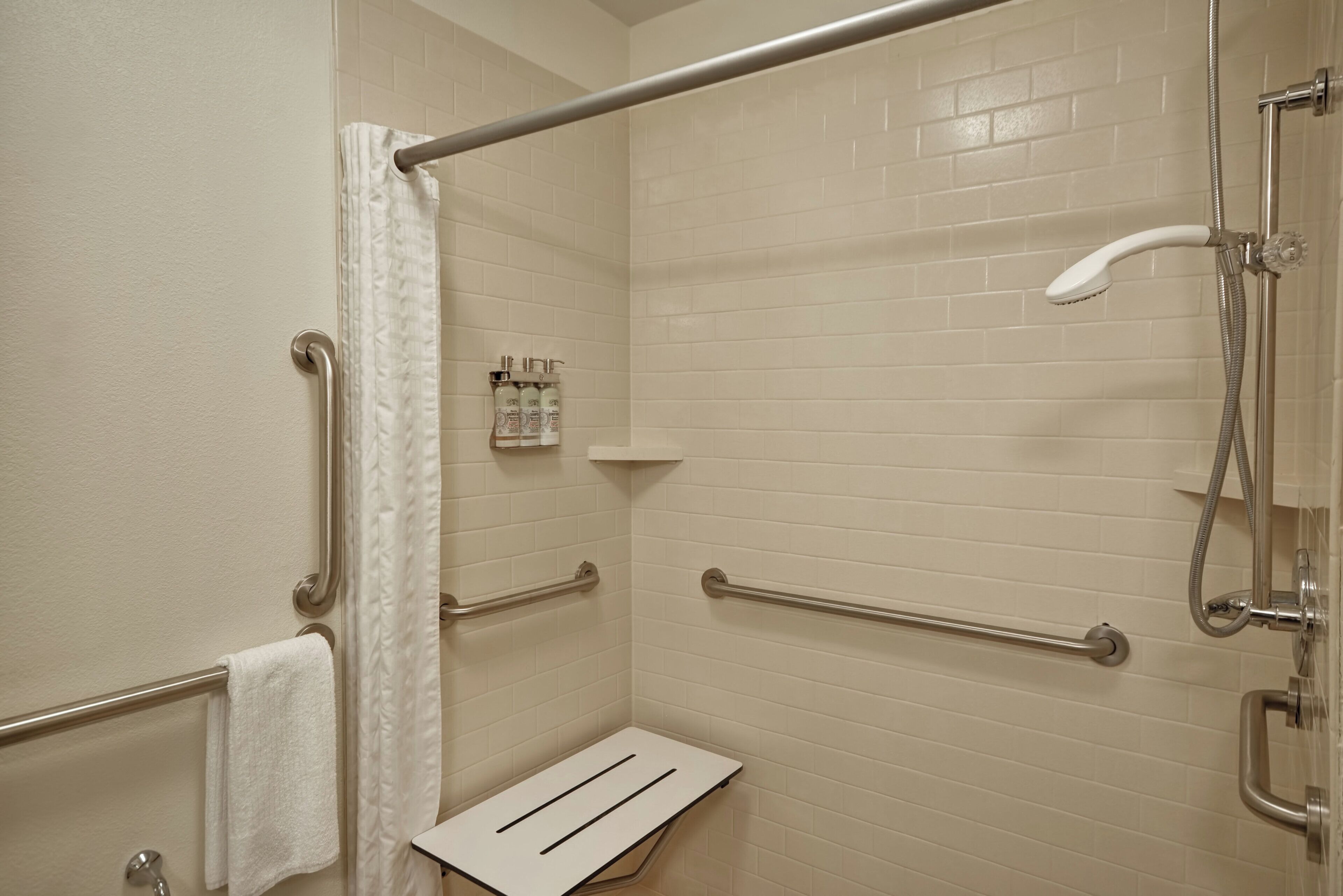 Une salle de bain avec des murs carrelés de carreaux blancs, une douche équipée d’un pommeau de douche à main et d’une barre d’appui.