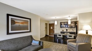 A modern hotel room with a gray sofa, a round coffee table, and a gray armchair. A kitchen area is visible in the background with a dining table and chairs. - Candlewood Suites Smyrna - Nashville by IHG (Smyrna)