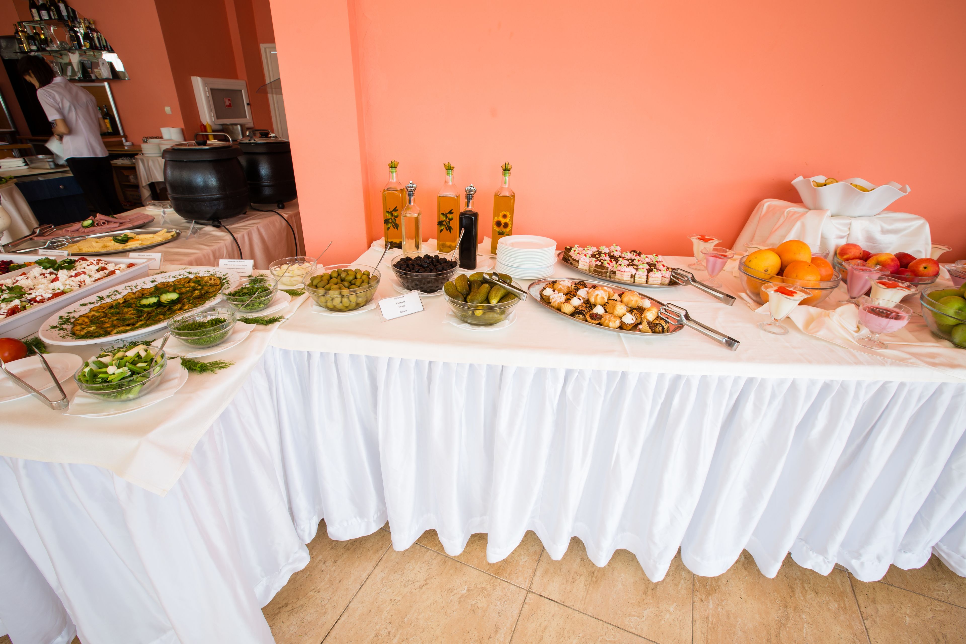 Mesa de buffet com diversos pratos e bebidas, área de cozinha ao fundo, e uma pessoa preparando comida.
