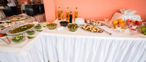 Mesa de buffet com diversos pratos e bebidas, área de cozinha ao fundo, e uma pessoa preparando comida.
