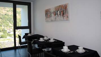 A dining area with tables set for four, each with white plates, cups, and cutlery. There is a large window providing a view of greenery and buildings.