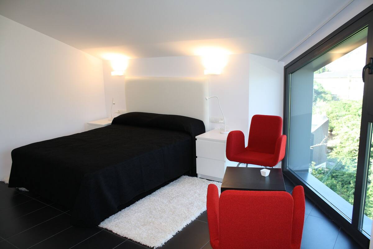 Een moderne slaapkamer met een zwart bed, een witte nachtkastje, een rode stoel en een groot raam met uitzicht op groen.