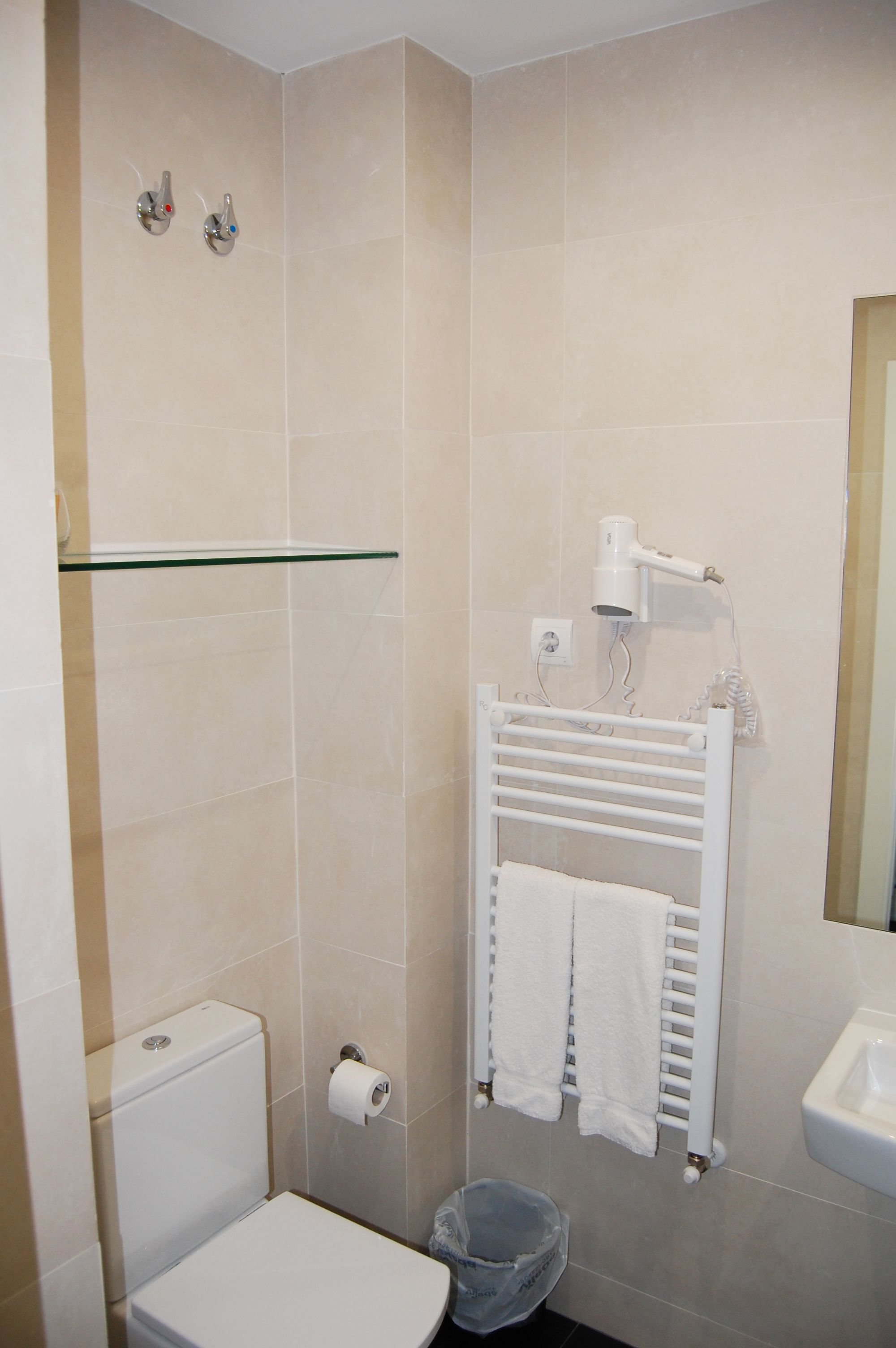 A bathroom with a toilet, a glass shelf, a towel rack with towels, a hairdryer, and a sink.
