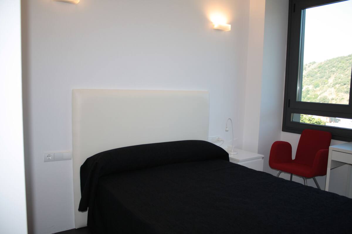 Een slaapkamer met een bed, een rode stoel, een bureau en een raam met uitzicht op een heuvel.