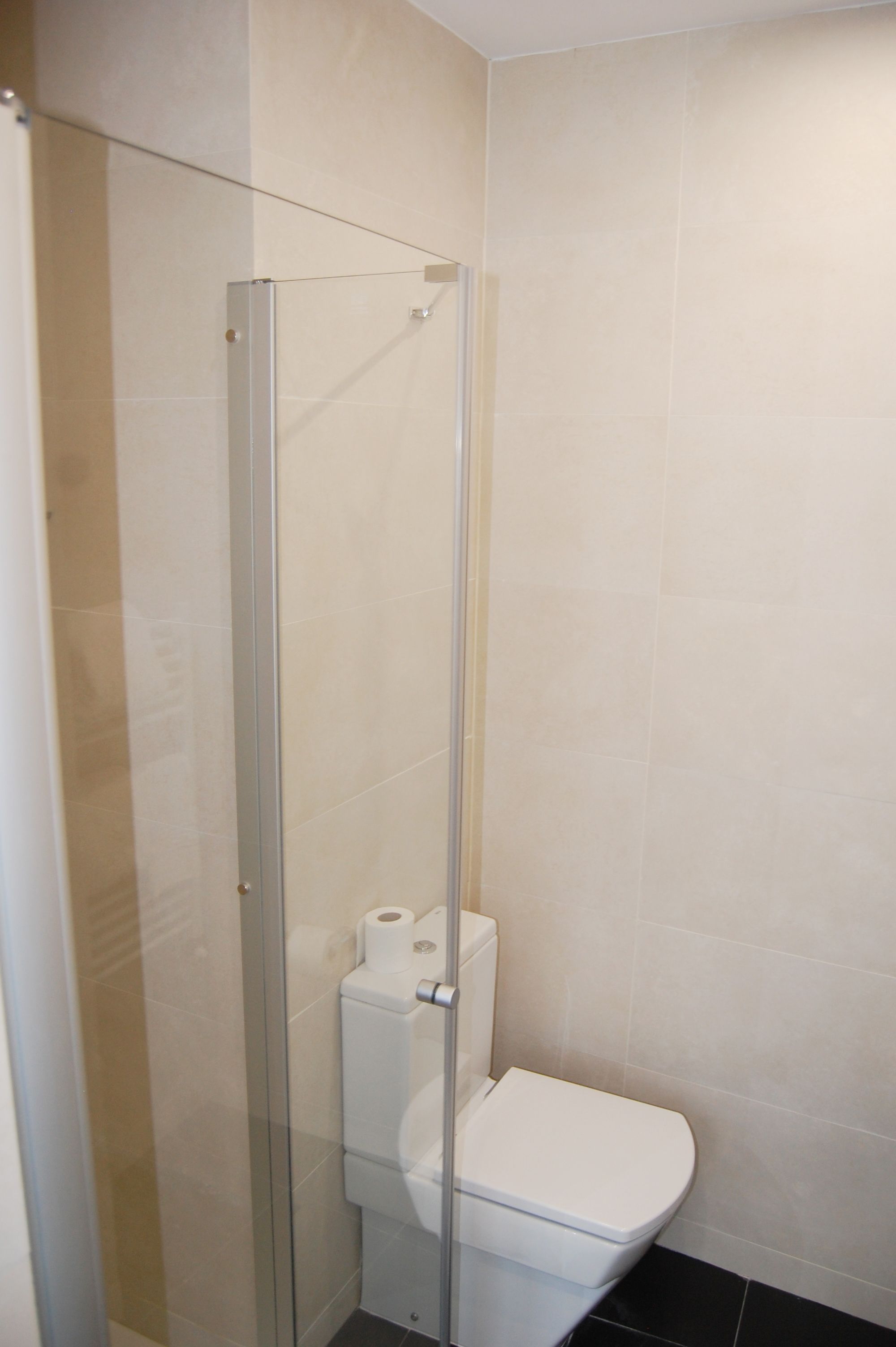 Un baño con ducha envuelta en vidrio, inodoro y paredes con azulejos color crema.