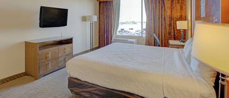 Ett hotellrum med en säng, en tv som är monterad på väggen, ett skrivbord med en lampa och ett fönster med gardiner.