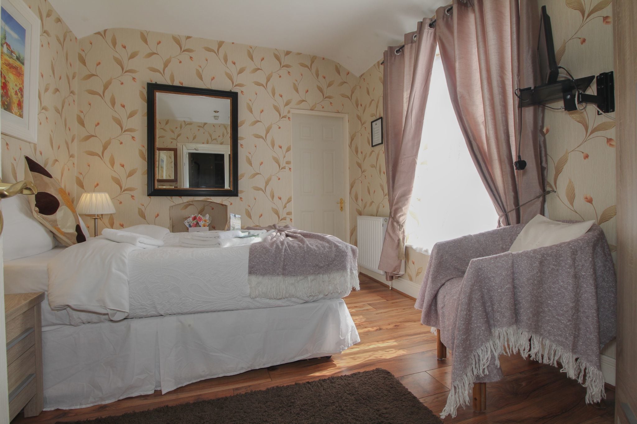 A bedroom with a double bed, a bedside table, a nightstand, a dresser, a mirror, a window with curtains, and a radiator.