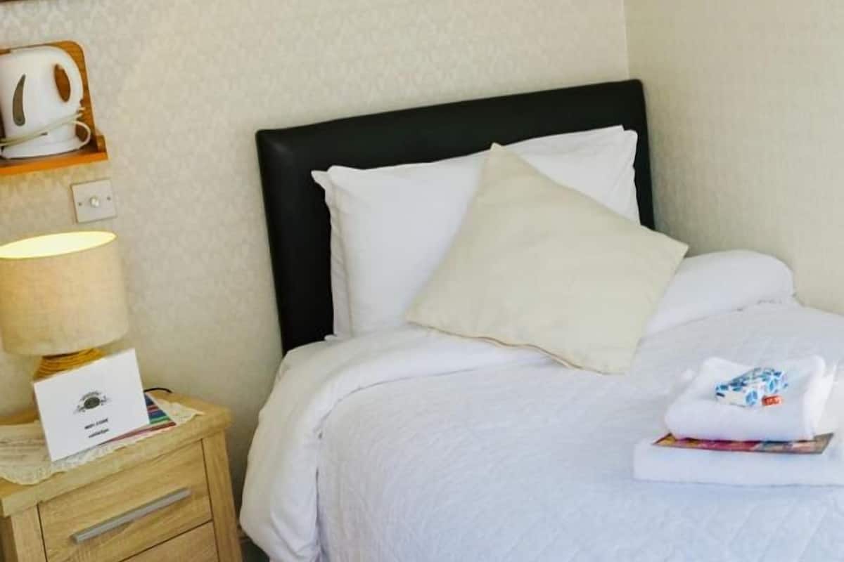 A hotel room with a bed, a bedside table with a lamp, a framed picture on the wall, and a small shelf with a kettle and a box.