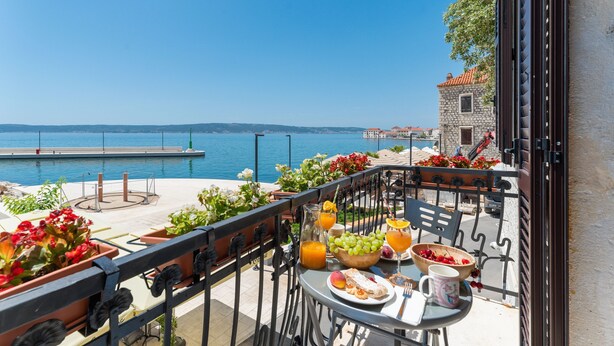 Un balcón con una mesa lista para el desayuno, con vistas a un cuerpo de agua y un edificio de piedra.