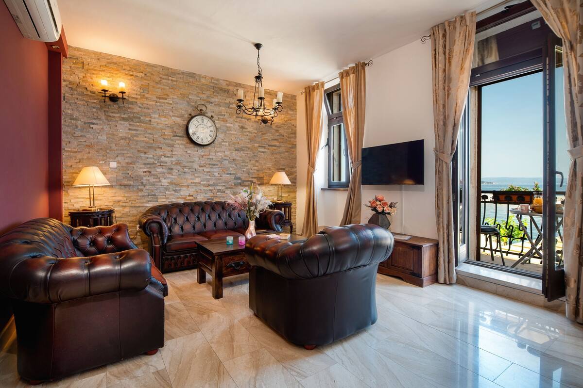 Una sala de estar con un sofá de cuero, una mesa de centro y un televisor montado en la pared. Hay un reloj en la pared y una ventana con cortinas.