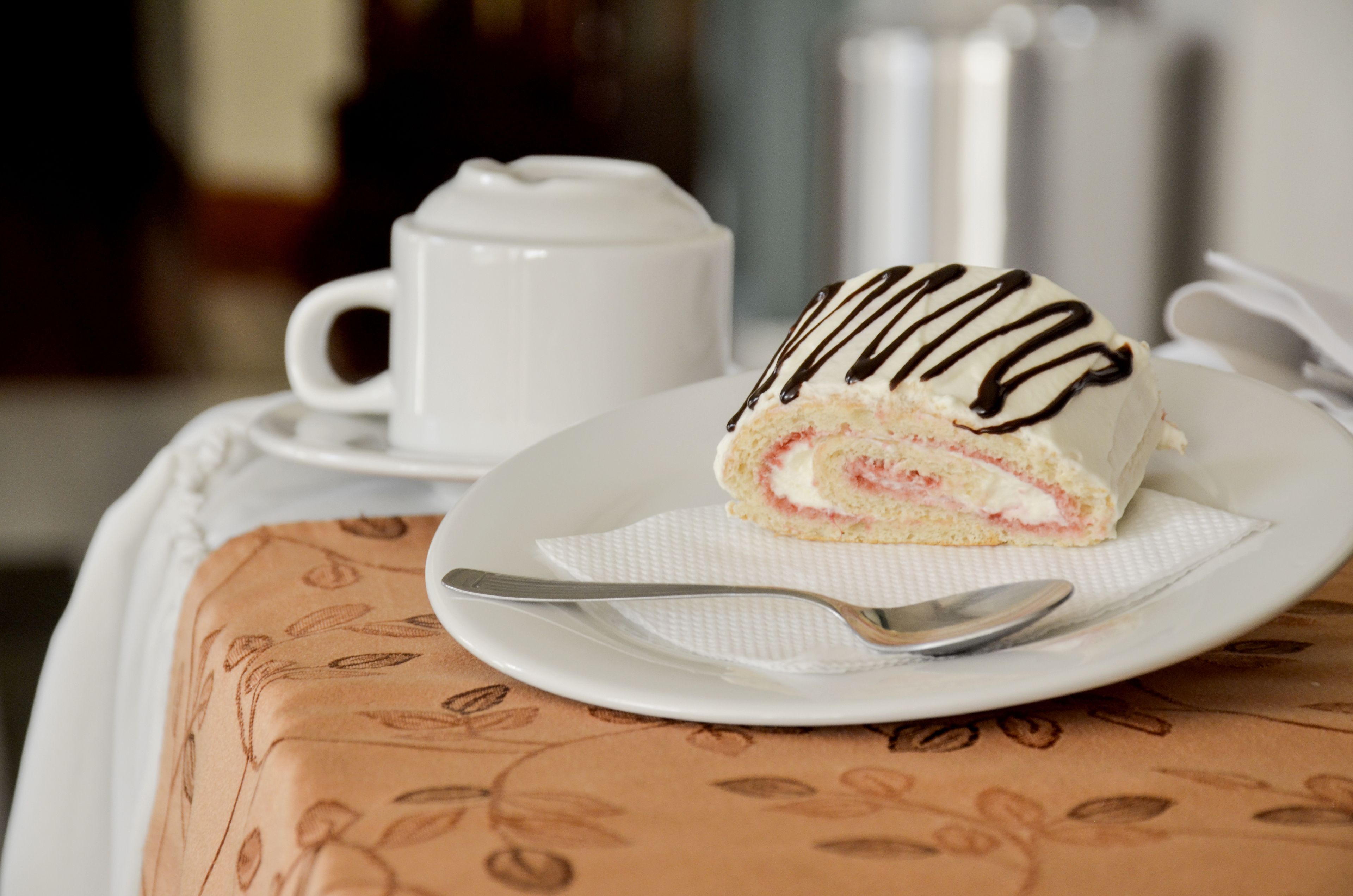 Una rebanada de pastel con glaseado de chocolate sobre un plato blanco, una cuchara y una taza blanca sobre una mesa con mantel estampado.