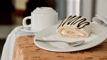 Una rebanada de pastel con glaseado de chocolate sobre un plato blanco, una cuchara y una taza blanca sobre una mesa con mantel estampado.