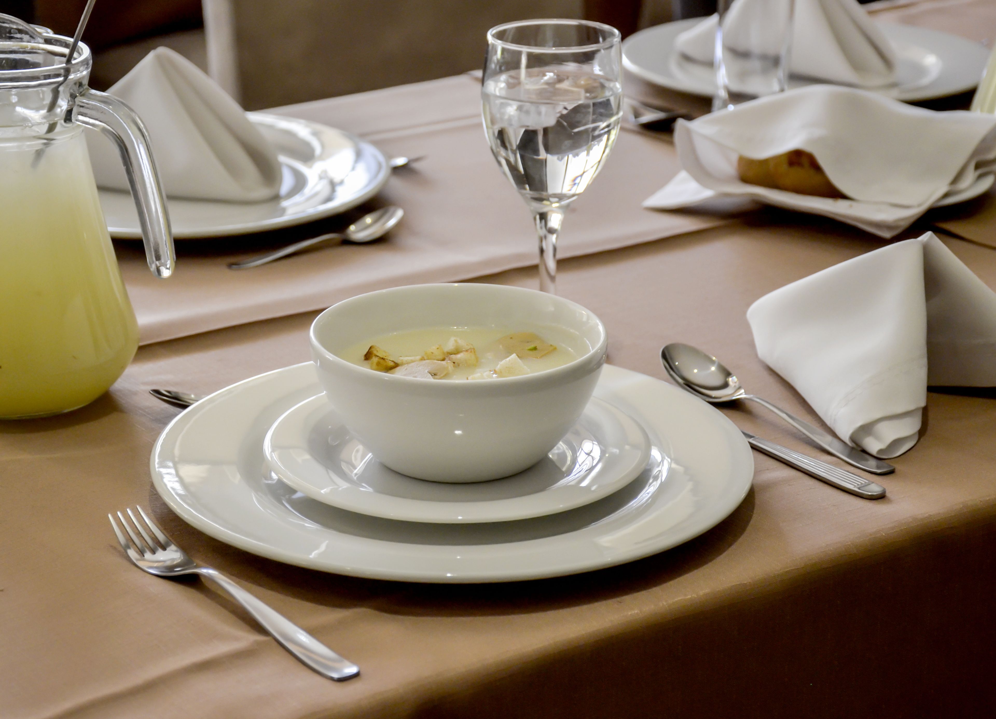 A dining table set with a bowl of soup, a glass of water, a pitcher of a yellow beverage, and cutlery.