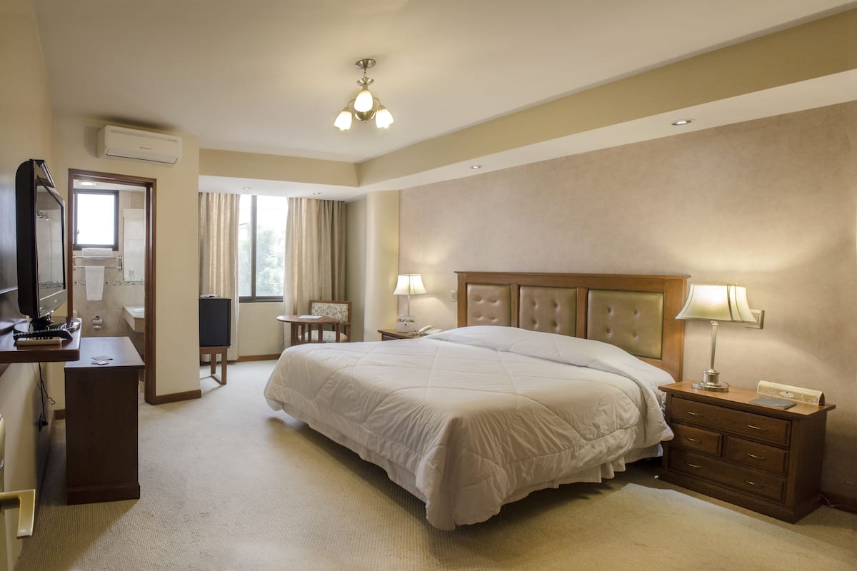A hotel room with a large bed, a desk, a chair, a desk lamp, and a window with curtains.