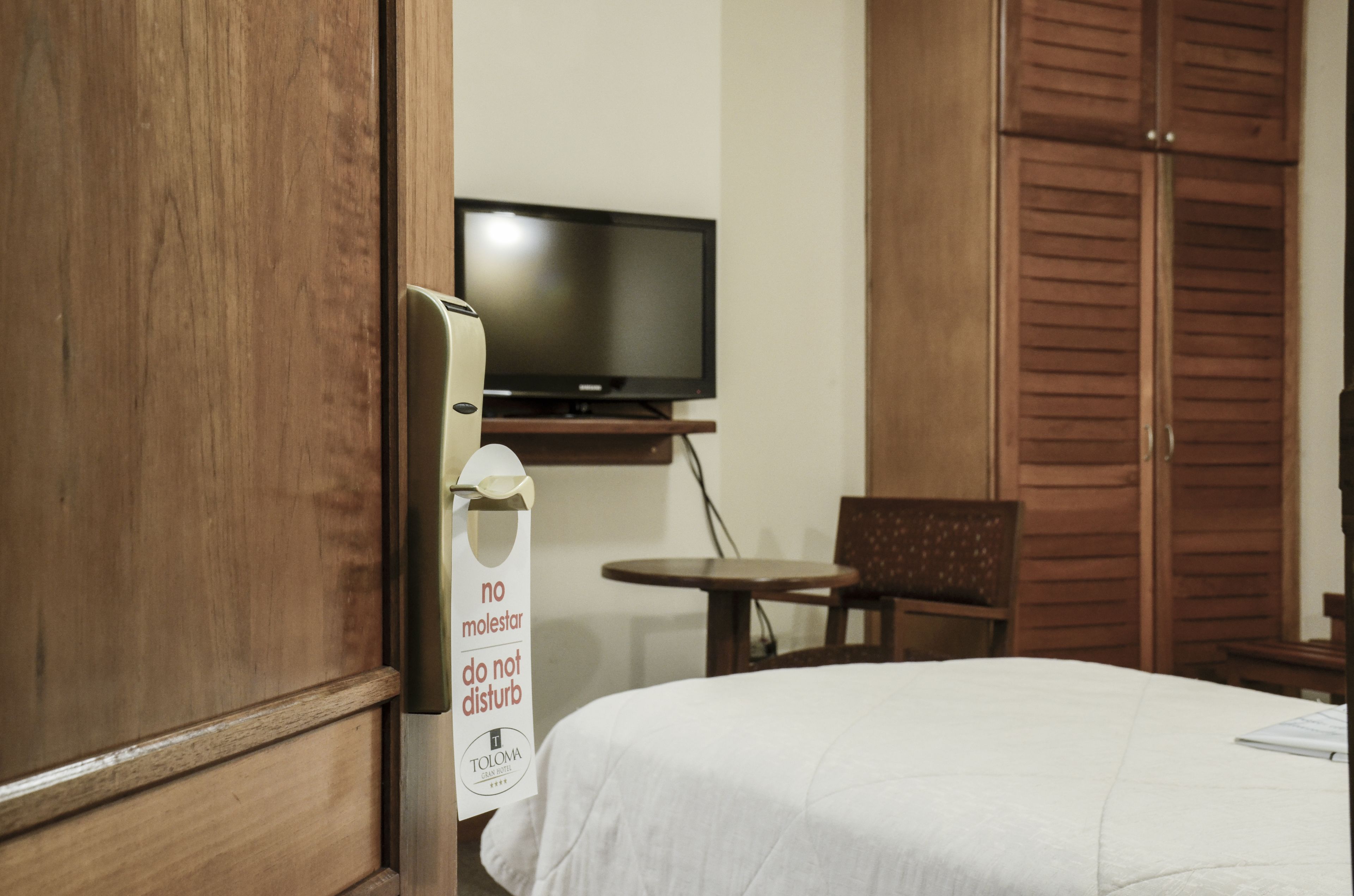 Een hotelkamer met een bed, een televisie op een standaard, een klein tafeltje en een stoel. Op de deur hangt een bordje met de tekst 'no molestar do not disturb'.