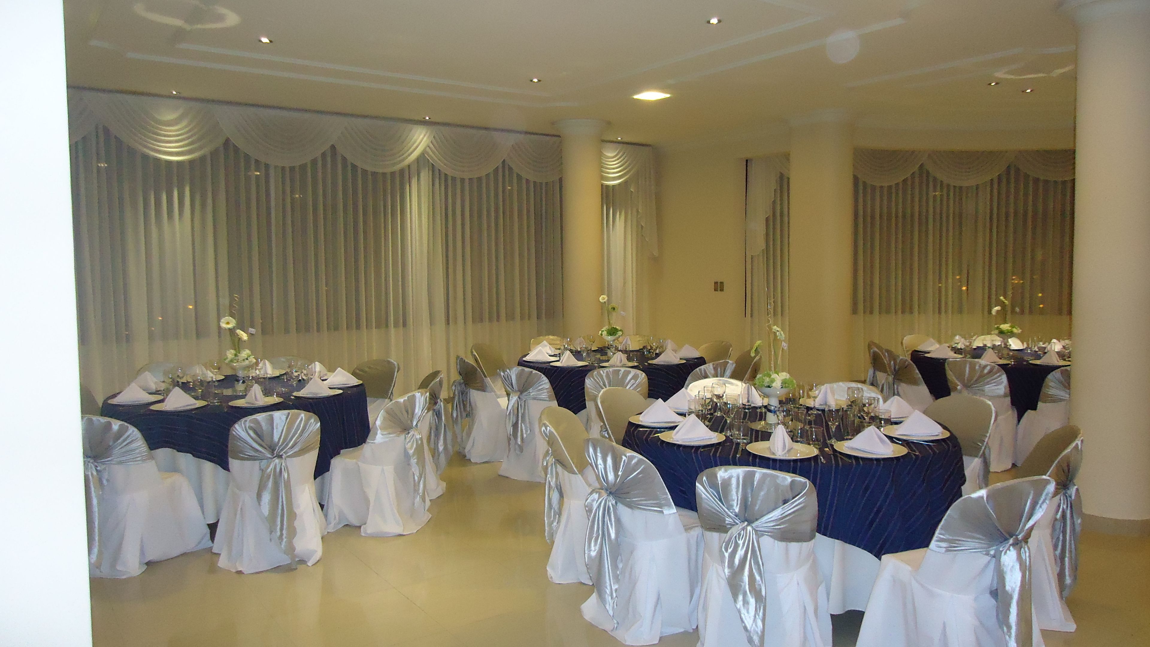 Un salón de banquetes con mesas redondas dispuestas para un evento formal, con manteles blancos y plateados, sillas con cintas de plata y centros de mesa florales.