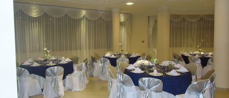 Un salón de banquetes con mesas redondas dispuestas para un evento formal, con manteles blancos y plateados, sillas con cintas de plata y centros de mesa florales.