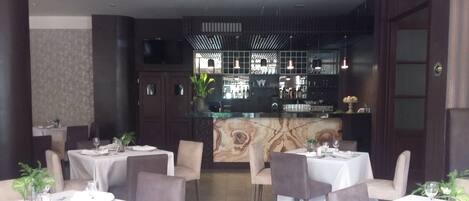Un restaurante con varias mesas, cada una con manteles blancos, platos y cubiertos. Hay plantas en macetas sobre las mesas, y la sala tiene un diseño moderno con paredes oscuras y un muro decorativo de mármol.