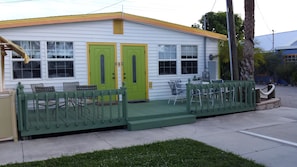 A white building with two green doors, a covered porch with chairs, and a small garden area.