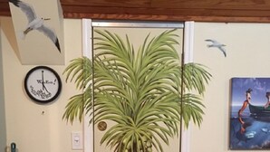 Quarto com porta adornada por um mural tropical, relógio de parede, quadro emoldurado, vassoura e lixeira.