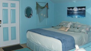 Quarto com cama, porta, espelho fixado na parede e uma pintura de uma paisagem marítima.