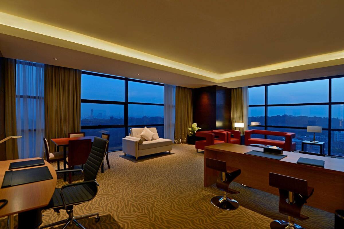 A hotel room with a large window, a desk, a sofa, and a red armchair.