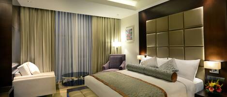A modern hotel room with a large bed, a seating area with a sofa and armchair, a small table, and a large window with curtains.