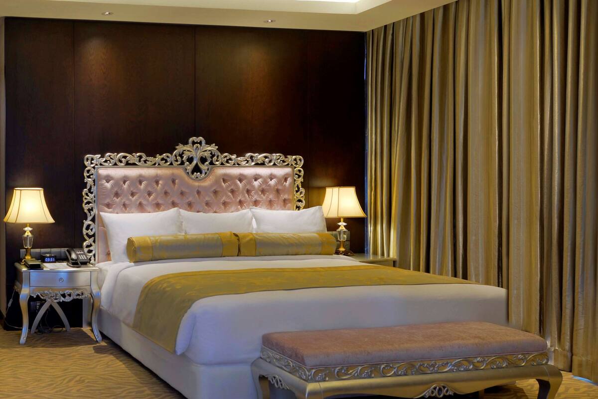 A luxurious hotel room with a large bed, ornate headboard, and a matching bench. There are two bedside tables with lamps and a telephone.