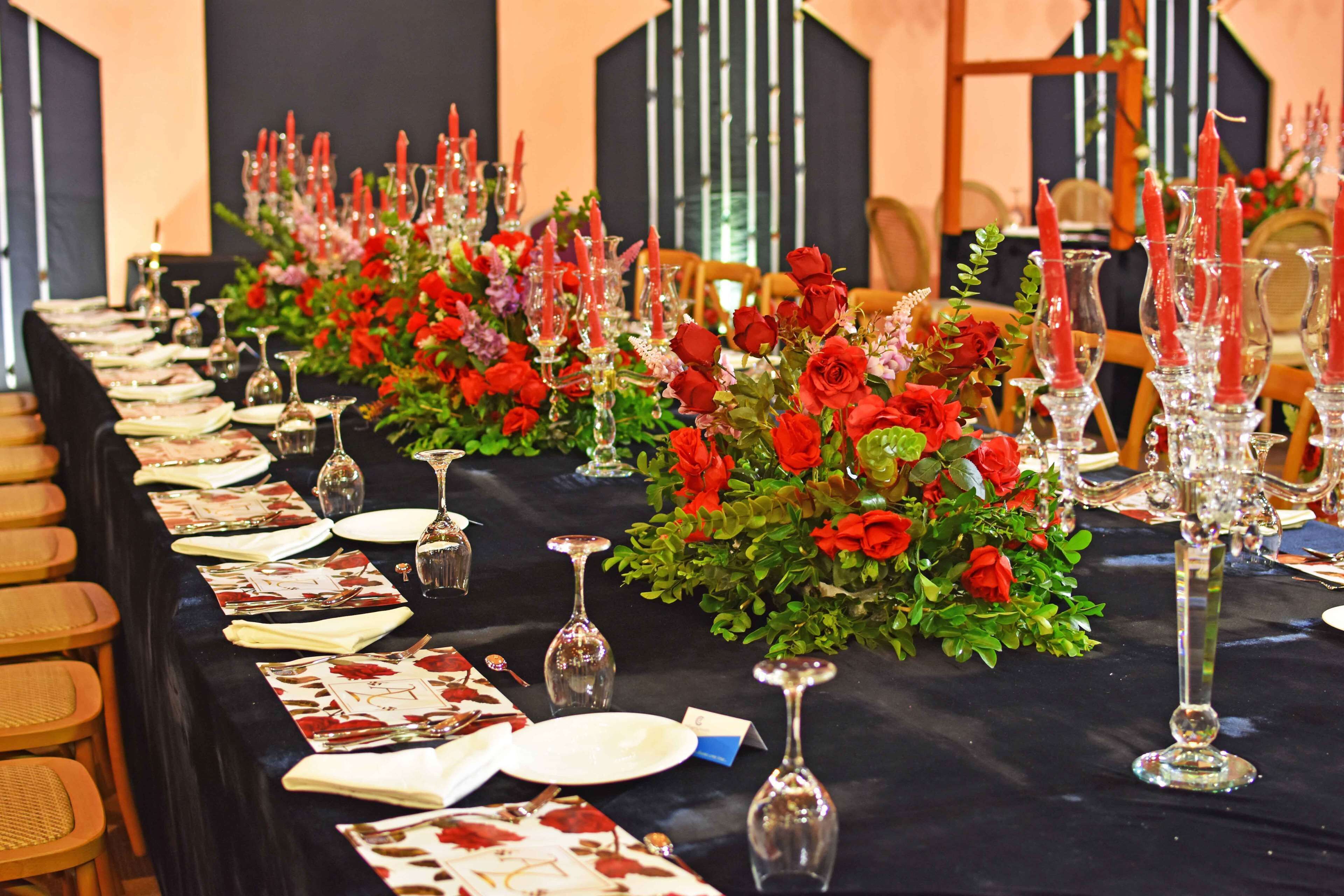A long dining table set with red roses, clear glassware, and neatly arranged plates and cutlery.