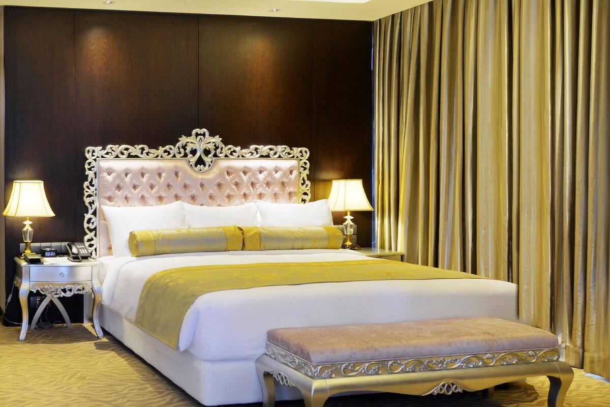 A large bed with a tufted headboard, two bedside tables with lamps, a bench, and a patterned carpet.