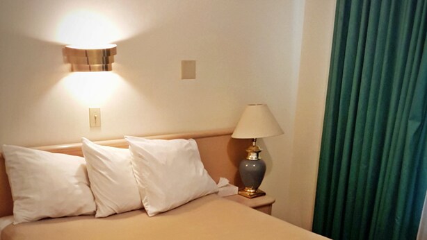 A neatly made bed with a beige bedspread and a floral-patterned skirt. There is a wall-mounted light fixture above the bed and a table lamp beside it. - Badlands Motel (Drumheller)