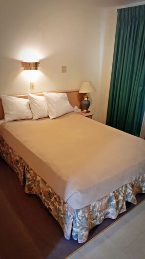 A neatly made bed with a beige bedspread and a floral-patterned skirt. There is a wall-mounted light fixture above the bed and a table lamp beside it. - Badlands Motel (Drumheller)