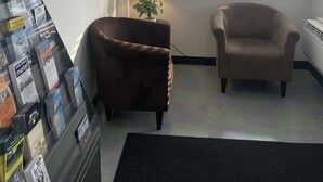 A room with two armchairs, a small table with a lamp, a bulletin board with travel brochures, and a window with blinds. - Bayside Inn  (St Ignace)