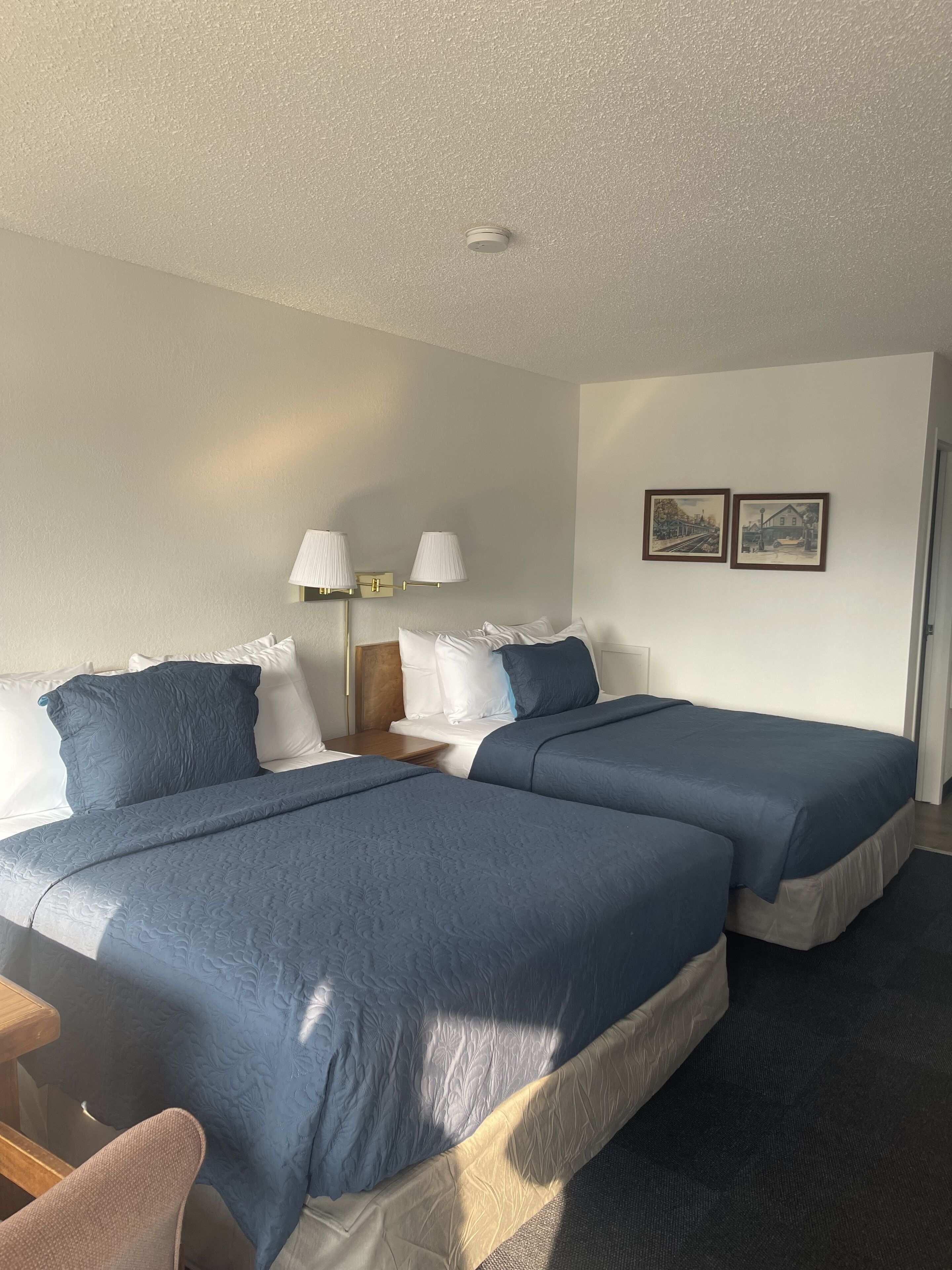 A hotel room with two beds, a nightstand, two wall-mounted lamps, and two framed pictures on the wall.