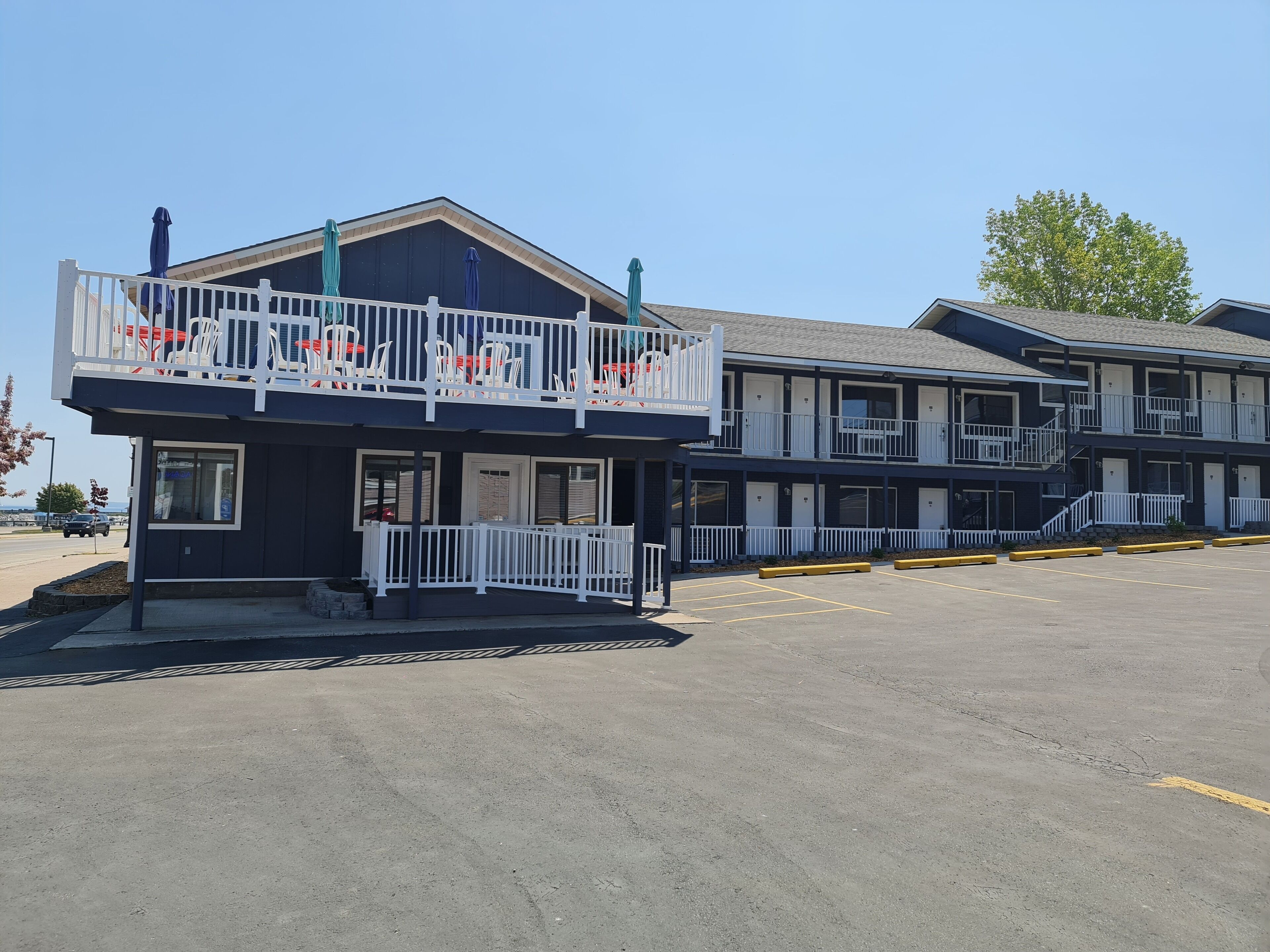 A two-story motel with a balcony on the ground floor, a parking lot in front, and a clear sky.