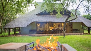 A thatched-roof building with an open dining area and a fire pit in the foreground. - Lokuthula Lodges (Victoria Falls)