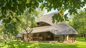 A thatched-roof house with an open porch and a dining area, surrounded by lush greenery. - Lokuthula Lodges (Victoria Falls)
