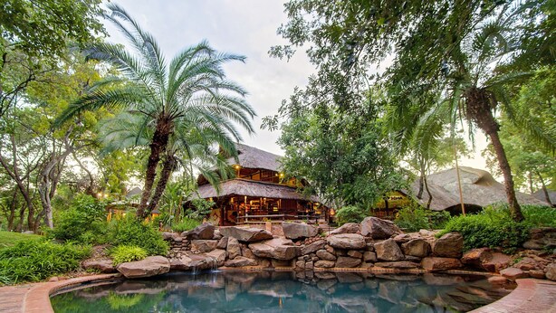 A pool surrounded by rocks and palm trees, with a building in the background. - Lokuthula Lodges (Victoria Falls)