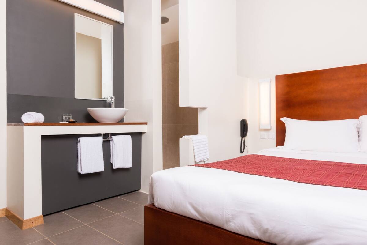 A hotel room with a bed, a wooden headboard, a white bedside table, a large mirror, a white sink, and a red bedspread.