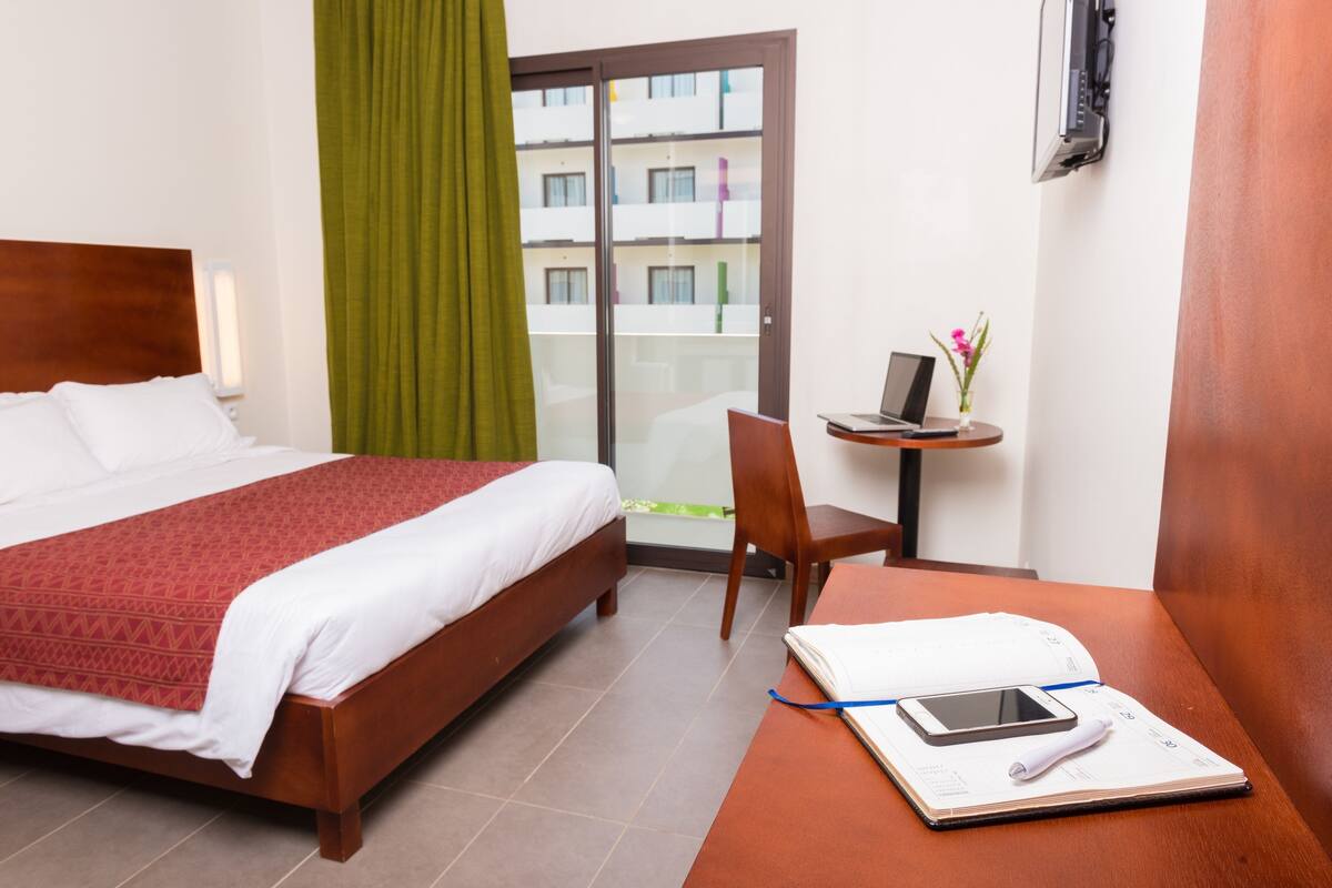 A hotel room with a bed, a desk with a laptop, a chair, and a window with a view of buildings.