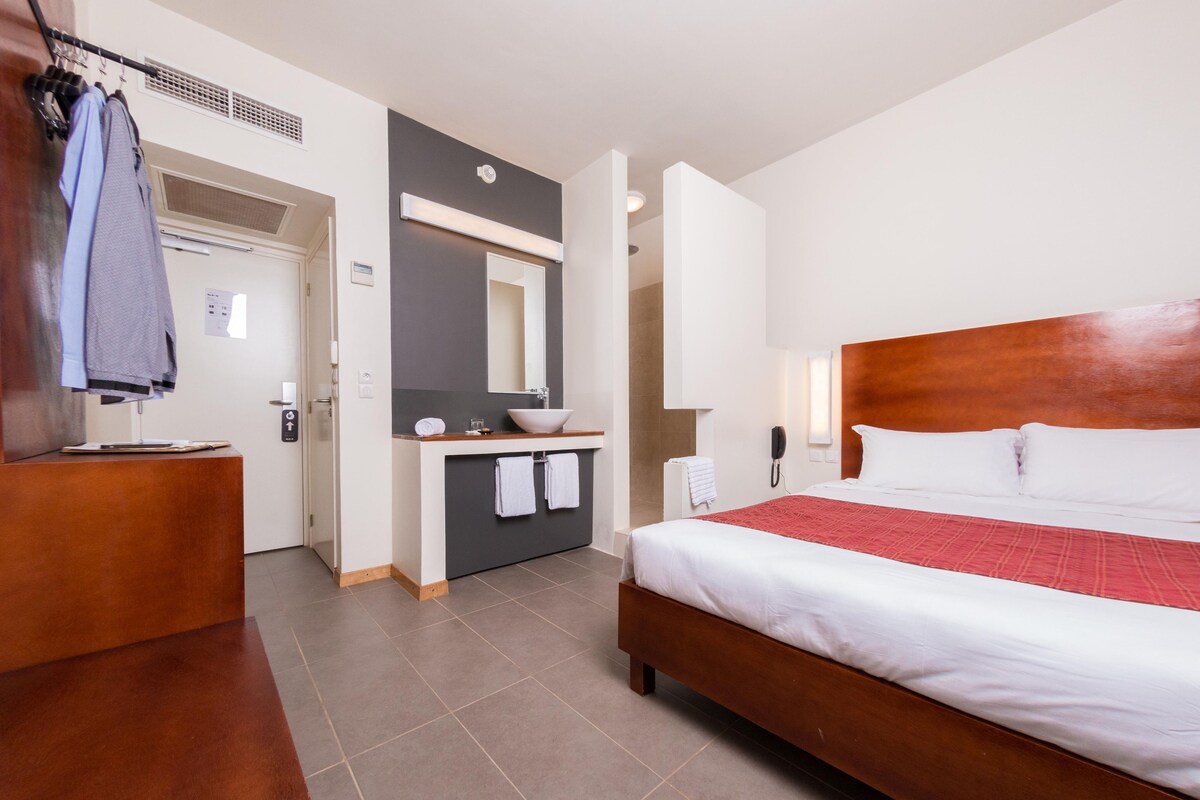 Habitación de hotel con cama de madera, mesita de noche, baño con lavabo y espejo, y perchero con ropa colgada.