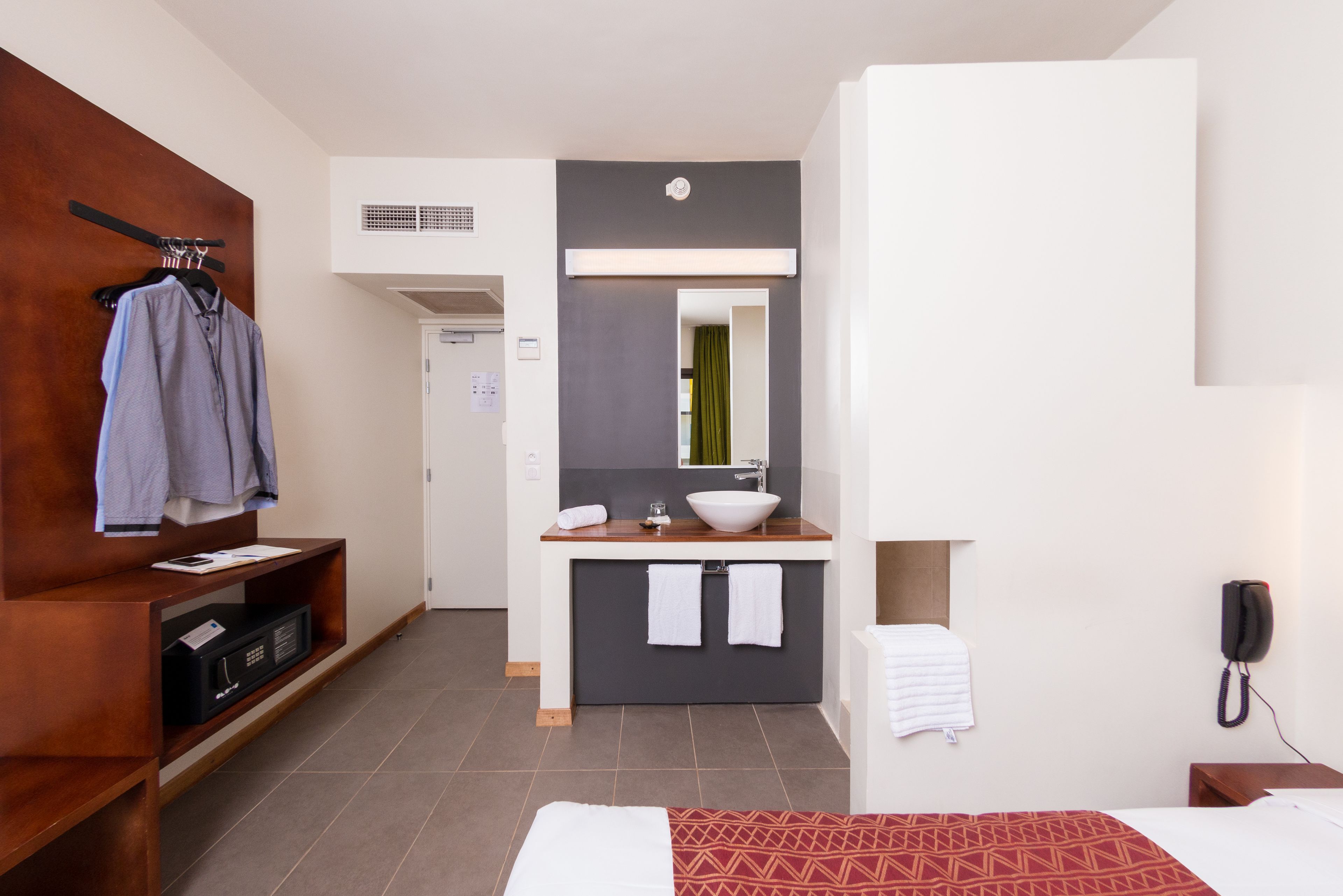 Une chambre d’hôtel comprenant un lit, une armoire en bois, une salle de bain avec un lavabo et un miroir, et un téléphone.
