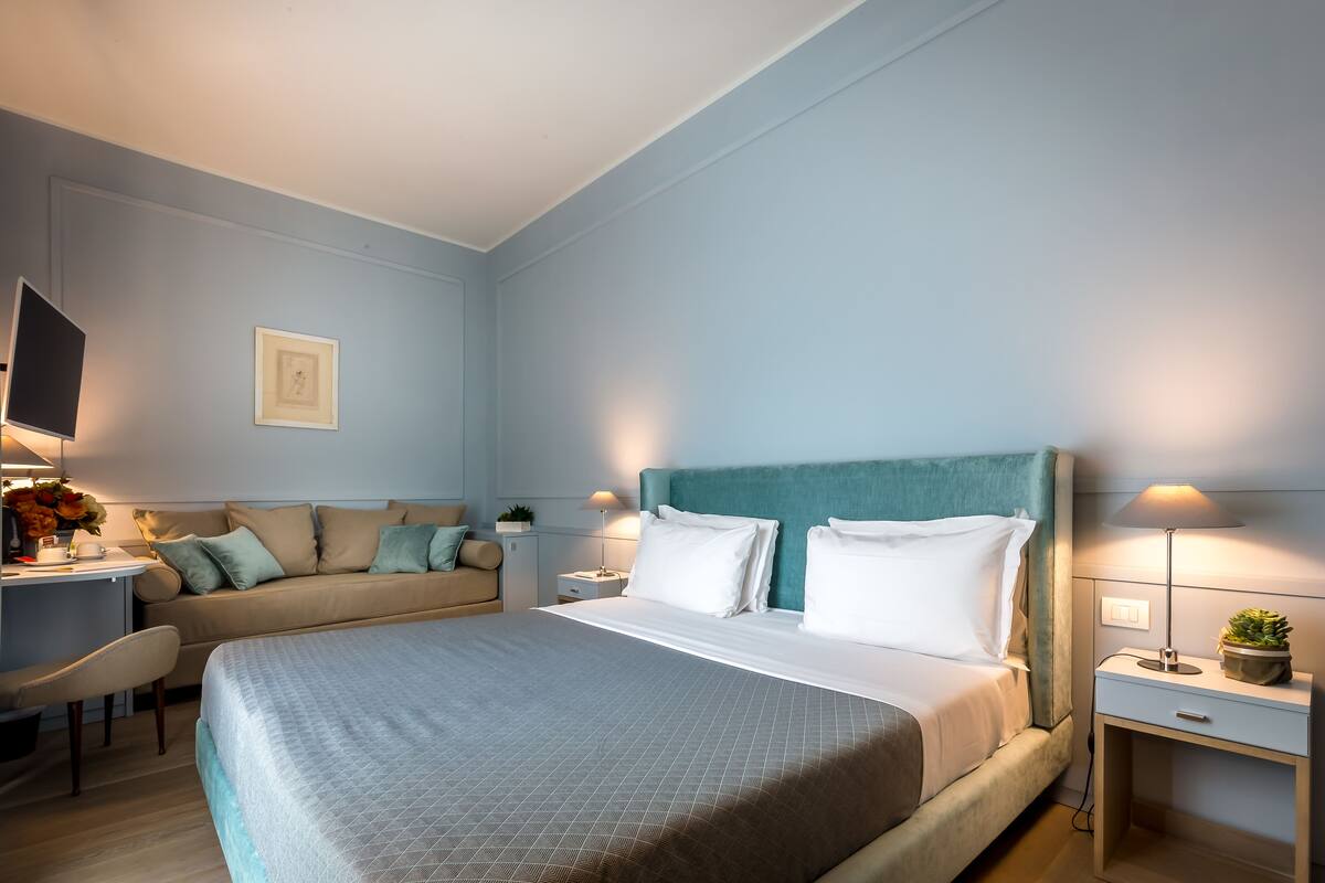 A modern hotel room with a large bed, a desk with a chair, a sofa, and a TV.