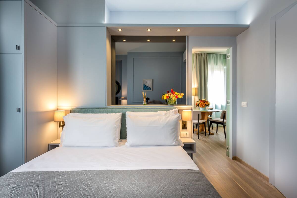 A hotel room with a large bed, bedside lamps, a mirror, a small table with a vase of flowers, and a view of the dining area.