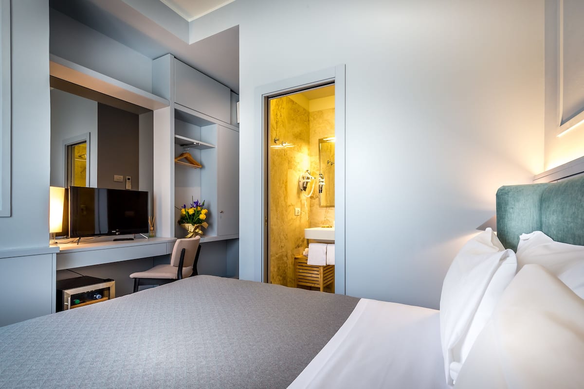A modern hotel room with a bed, a desk, a chair, a TV, and a bathroom visible through an open door.