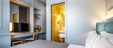 A modern hotel room with a bed, a desk, a chair, a TV, and a bathroom visible through an open door.